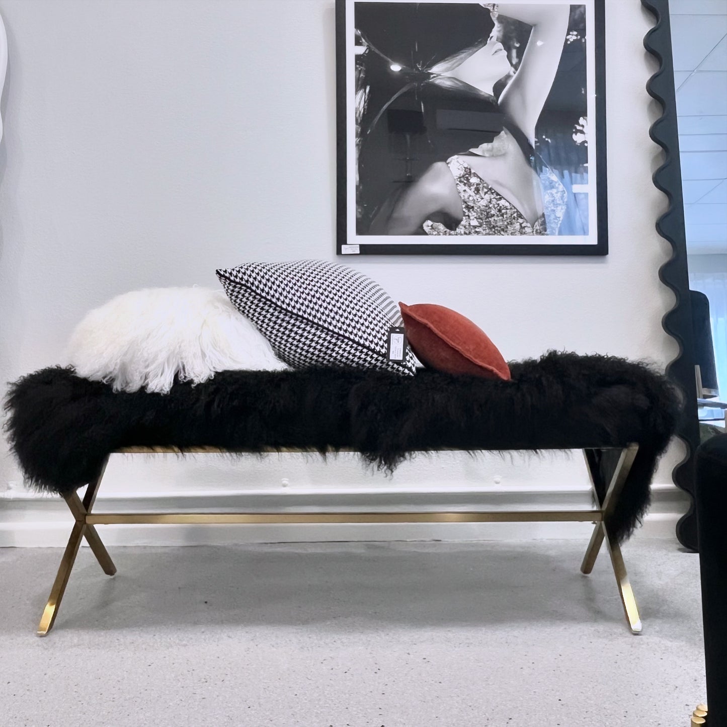 A black fur bench with gold finished legs, faux fur upholstery, and a brushed steel base, displayed in a showroom setting with decorative pillows on it.
