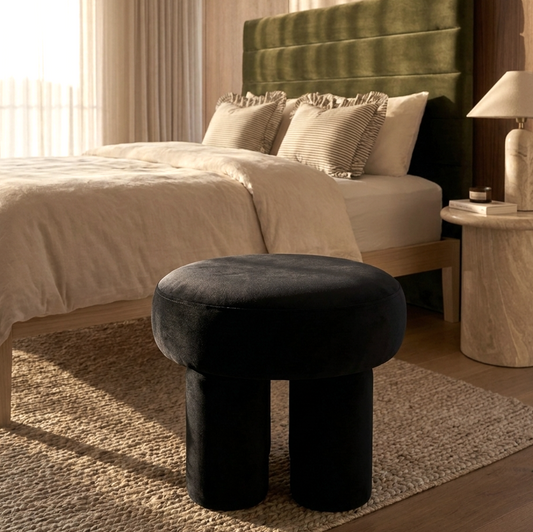 Black velvet stool in front of a bed with green headboard and beige bedding.