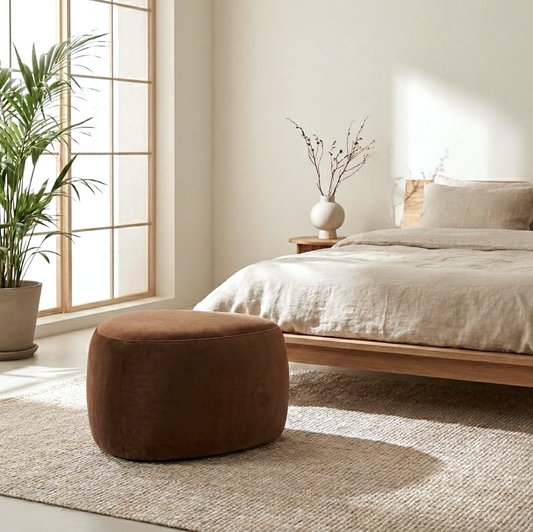 Cove pebble ottoman in cinnamon, rounded sculptural form in warm brown fabric styled at the end of a bed beside a palm plant