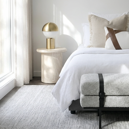 Nobu natural stone side table styled in a bedroom beside a bed with white linen and a warm gold pendant lamp