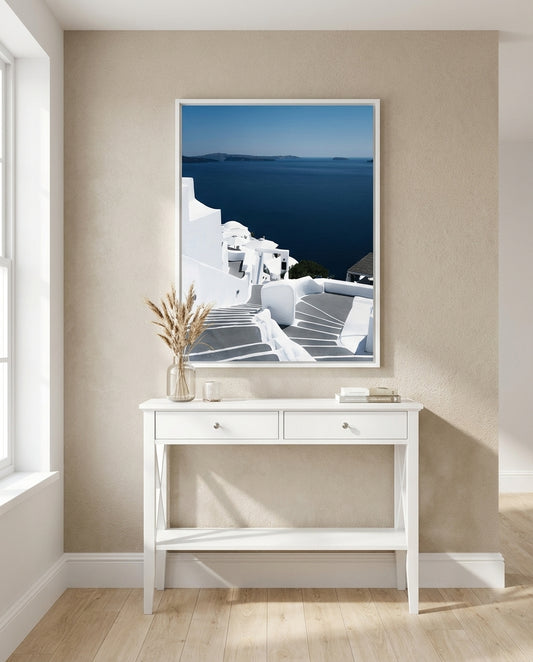 Framed artwork of a coastal scene above a white console table in a room with a window.