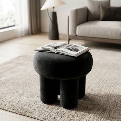 Interior Ave Black Ottoman Bentley - A round black velvet ottoman stool with cylindrical legs in a modern living room setting, with an open magazine resting on top.