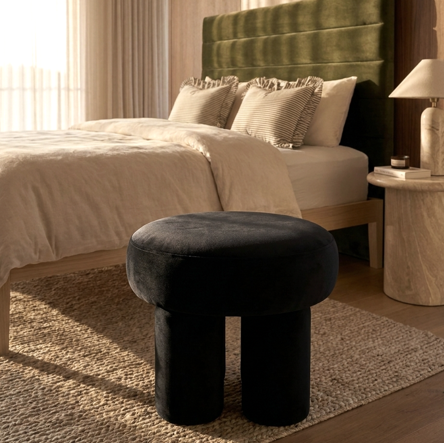 Black velvet stool in front of a bed with green headboard and beige bedding.