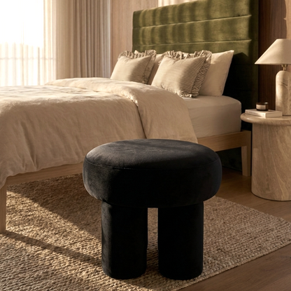 Black velvet stool in front of a bed with green headboard and beige bedding.