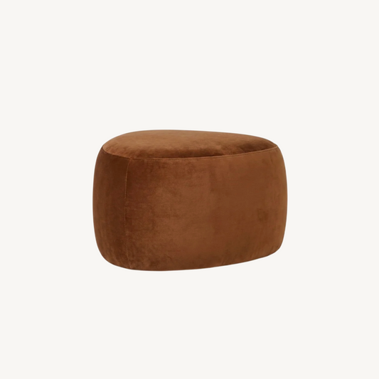 Cove pebble ottoman in cinnamon, rounded sculptural form in warm brown fabric styled at the end of a bed beside a palm plant