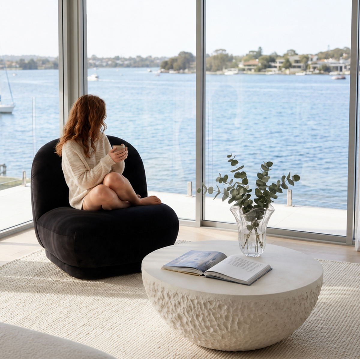 Lunar fossil stone coffee table with a woman sitting beside it in a dark textured chair, water views and a book on the table surface