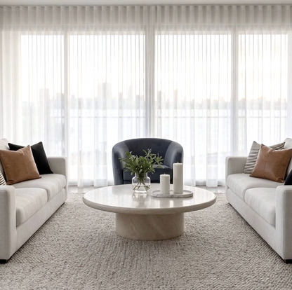A modern living room setup featuring two white sofas with brown and black accent pillows, a dark blue armchair, a round white marble coffee table, and a textured light gray rug in front of large white sheer curtains.