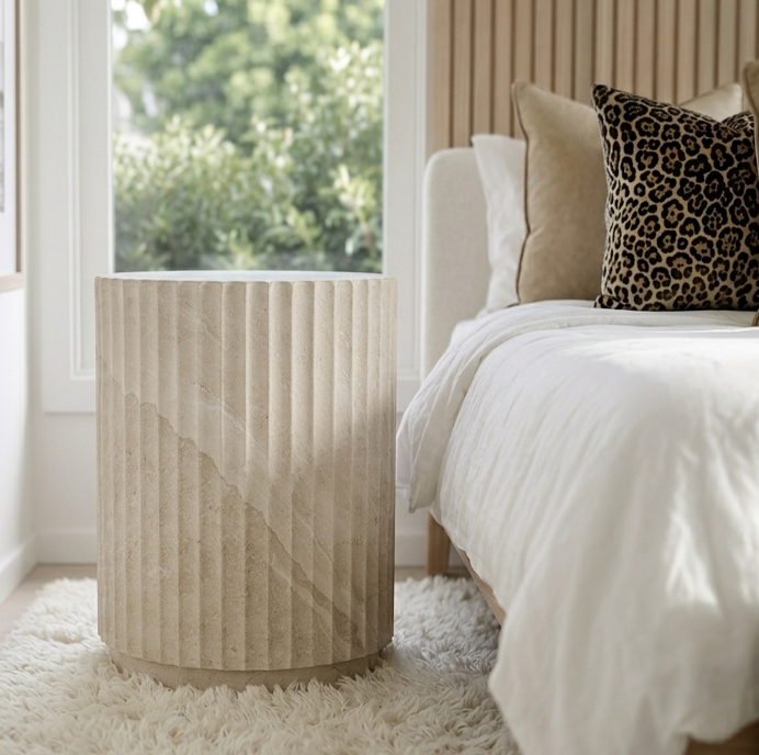 Vera fluted stone side table in a bright bedroom with white bedding and dark cushions beside a garden-view window