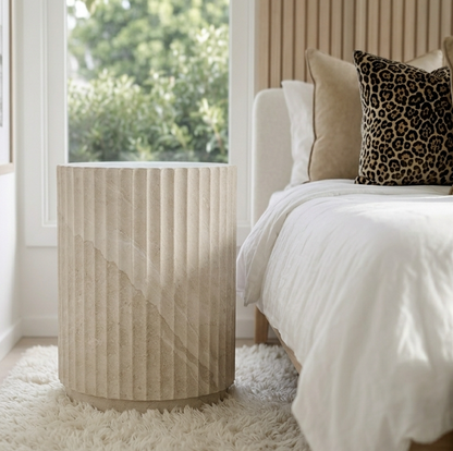 Vera fluted stone side table in a bright bedroom with white bedding and dark cushions beside a garden-view window
