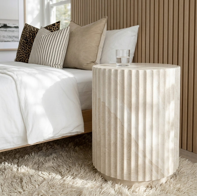 Vera fluted stone side table styled in a warm bedroom beside a bed with white and caramel linen and slatted timber headboard
