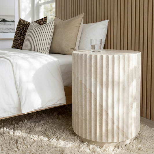 Vera fluted stone side table styled in a warm bedroom beside a bed with white and caramel linen and slatted timber headboard